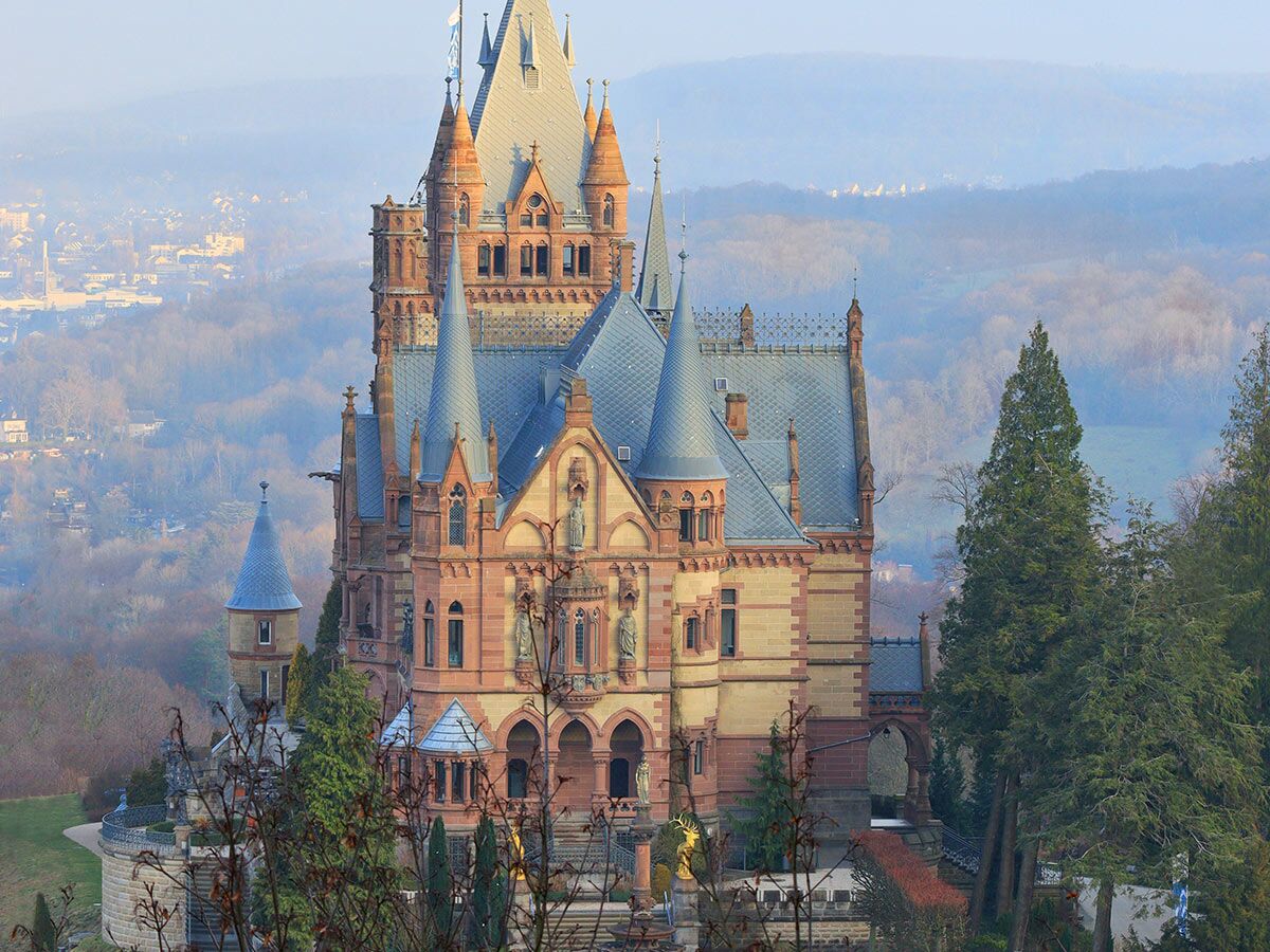 Schloss Drachenburg, ein imposantes historisches Bauwerk auf dem Drachenfels, mit roten Steinfassaden und grauen Türmen, umgeben von Bäumen und nebliger Landschaft.