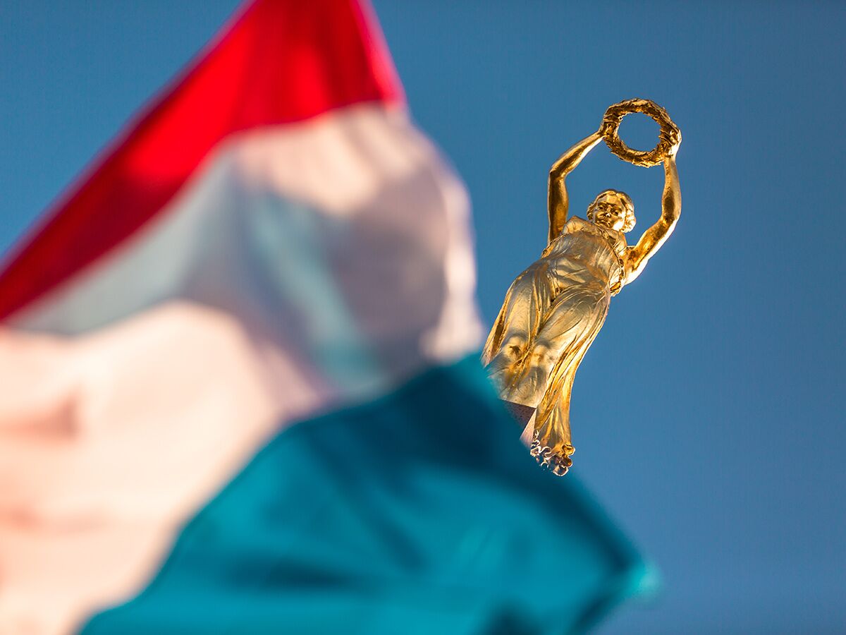 Goldene Gëlle Fra Statue, Wahrzeichen Luxemburgs, vor blauem Himmel mit wehender Nationalflagge im Vordergrund.
