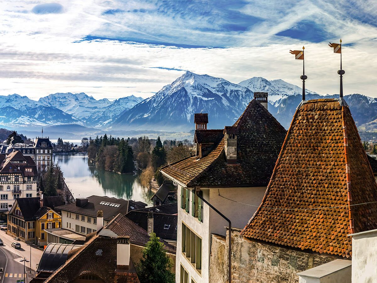Malerische Schweizer Stadt am Fluss mit historischen Gebäuden, ziegelgedeckten Dächern und schneebedeckten Alpen im Hintergrund.