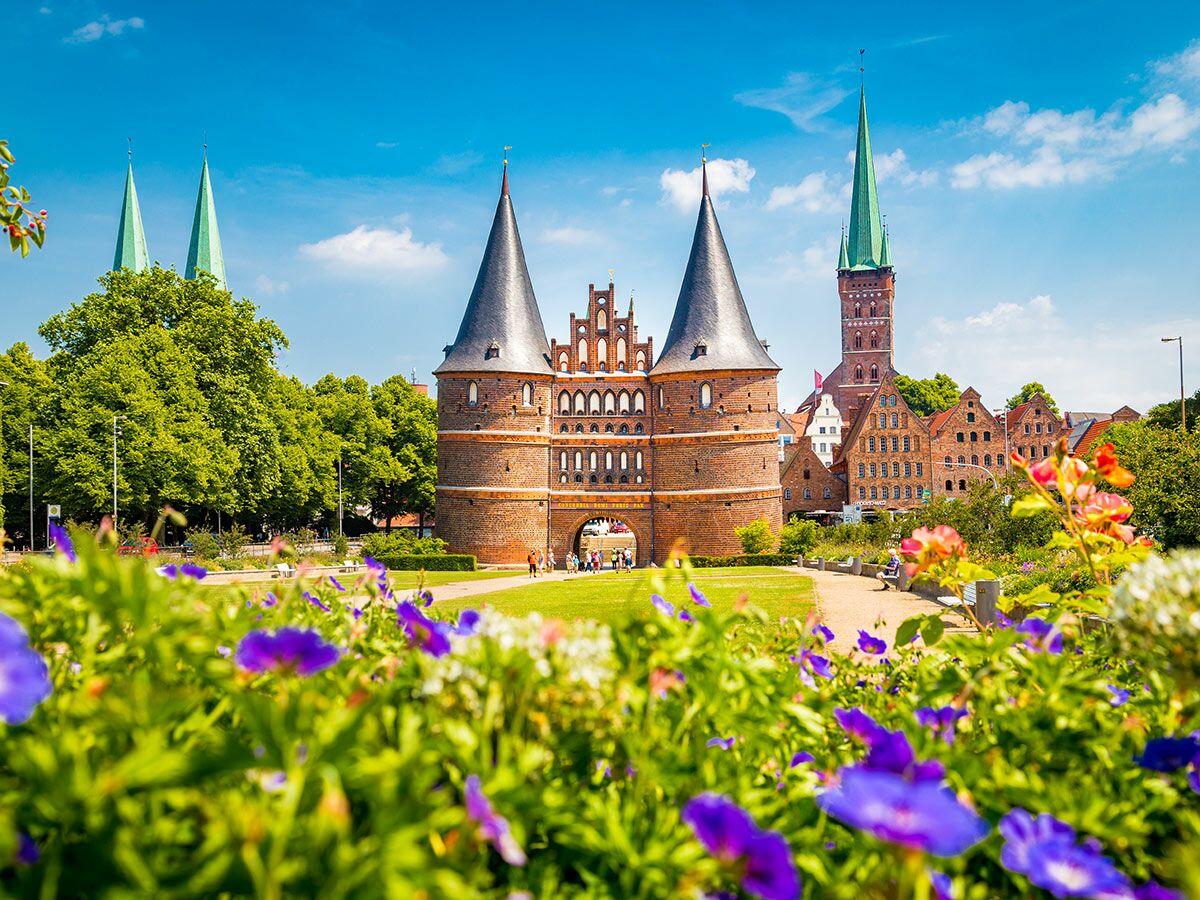 Das historische Holstentor in Lübeck mit Altstadtgebäuden, grünen Bäumen und farbigen Blumen unter blauem Himmel an einem Sommertag.