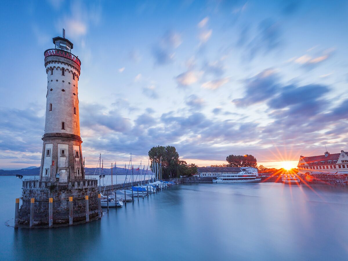 Der Neue Lindauer Leuchtturm im Hafen von Lindau am Bodensee bei Sonnenuntergang mit Booten und Wolkenhimmel.