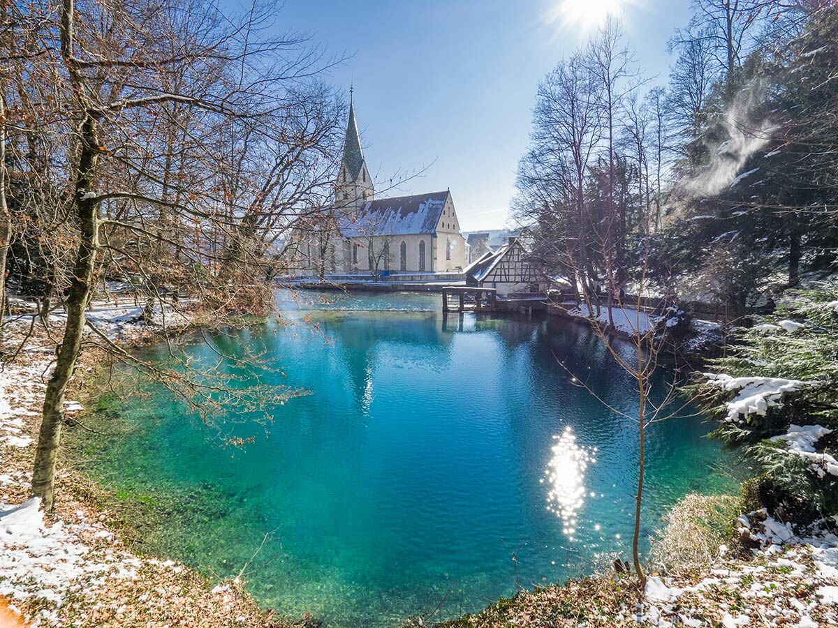 Blick auf einen leuchtend türkisen See mit schneebedeckter Uferlandschaft und einer Kirche im Hintergrund an einem sonnigen Wintertag.