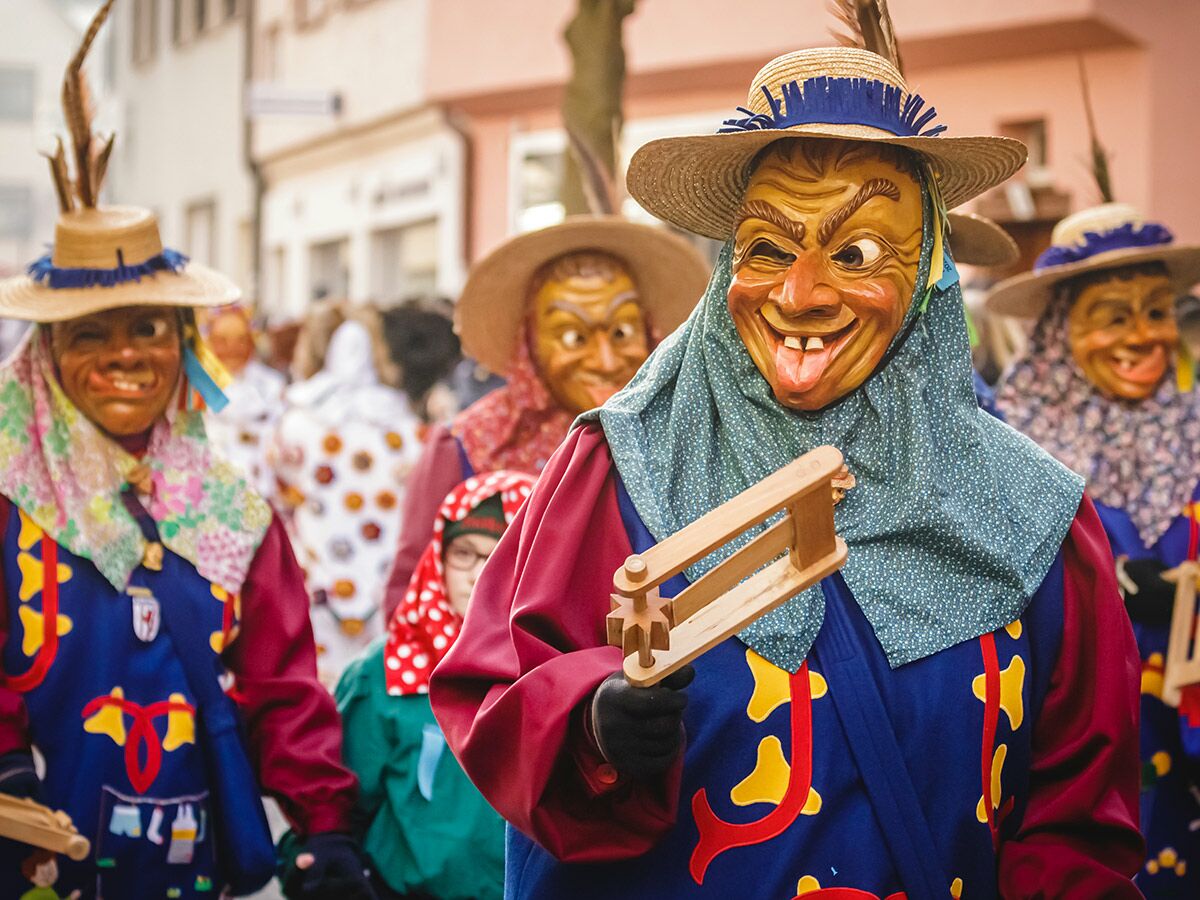 Karnevalisten mit traditionellen Holzmasken, Strohhüten und bunten Gewändern im Fasnetsumzug, einer zwinkert und streckt Zunge heraus.