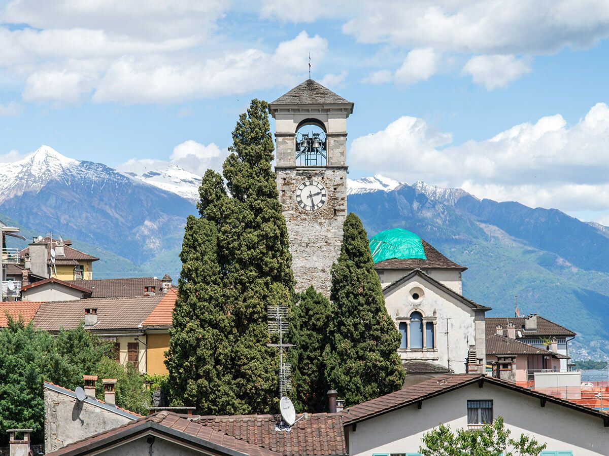 Steinerner Glockenturm mit Uhr und Glocken, umgeben von hohen Zypressen, vor schneebedeckten Bergen und Dächern.