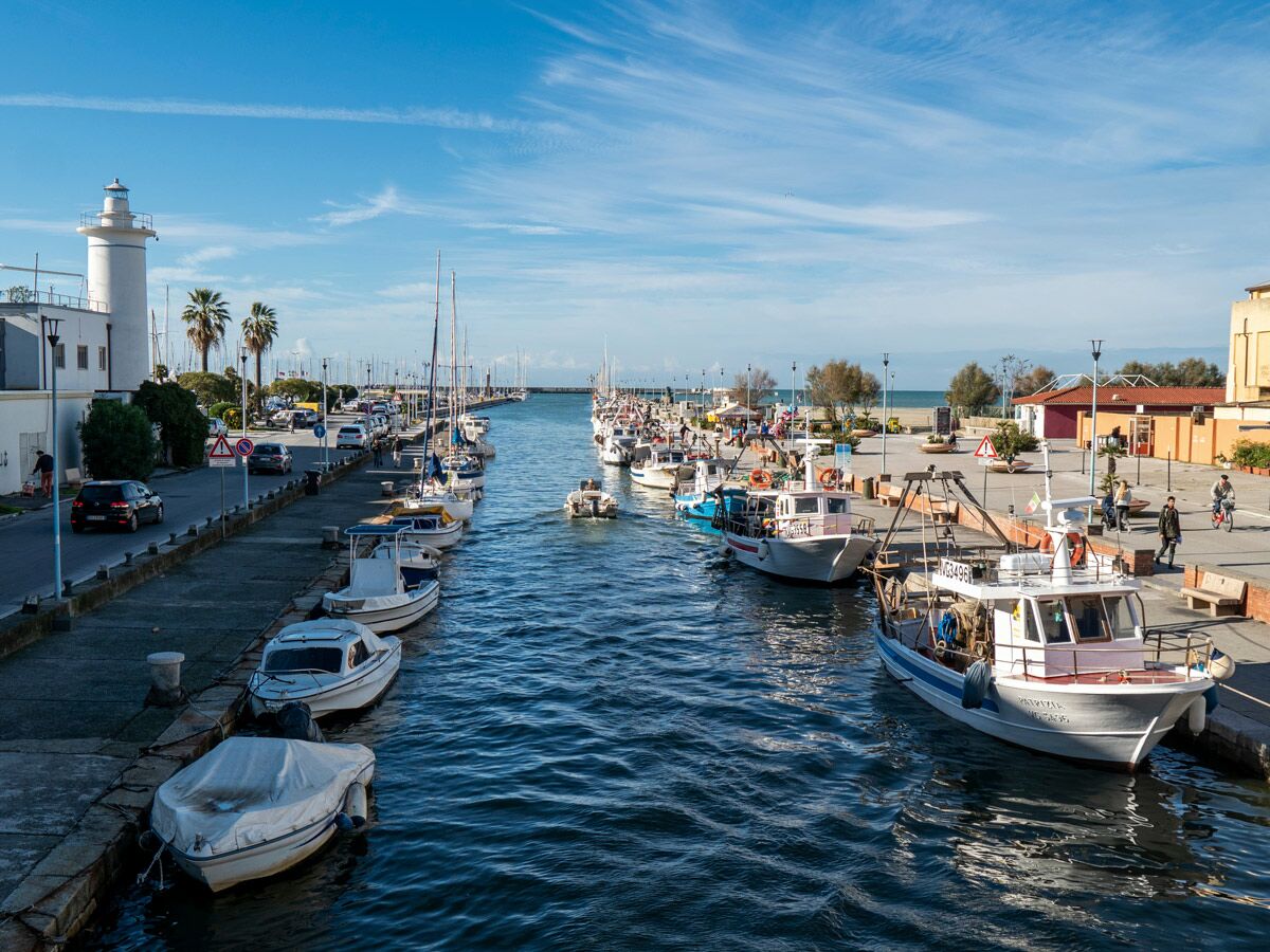 Blick auf einen belebten Hafenkanal mit zahlreichen Fischerbooten und Segelbooten, einem weißen Leuchtturm und einer Promenade mit Menschen bei Sonnenschein.
