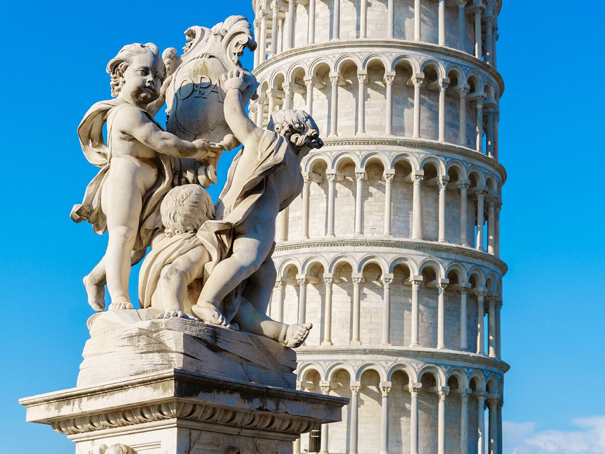 Marmorstatue von Cherubim mit Wappenschild vor dem Schiefen Turm von Pisa unter blauem Himmel.