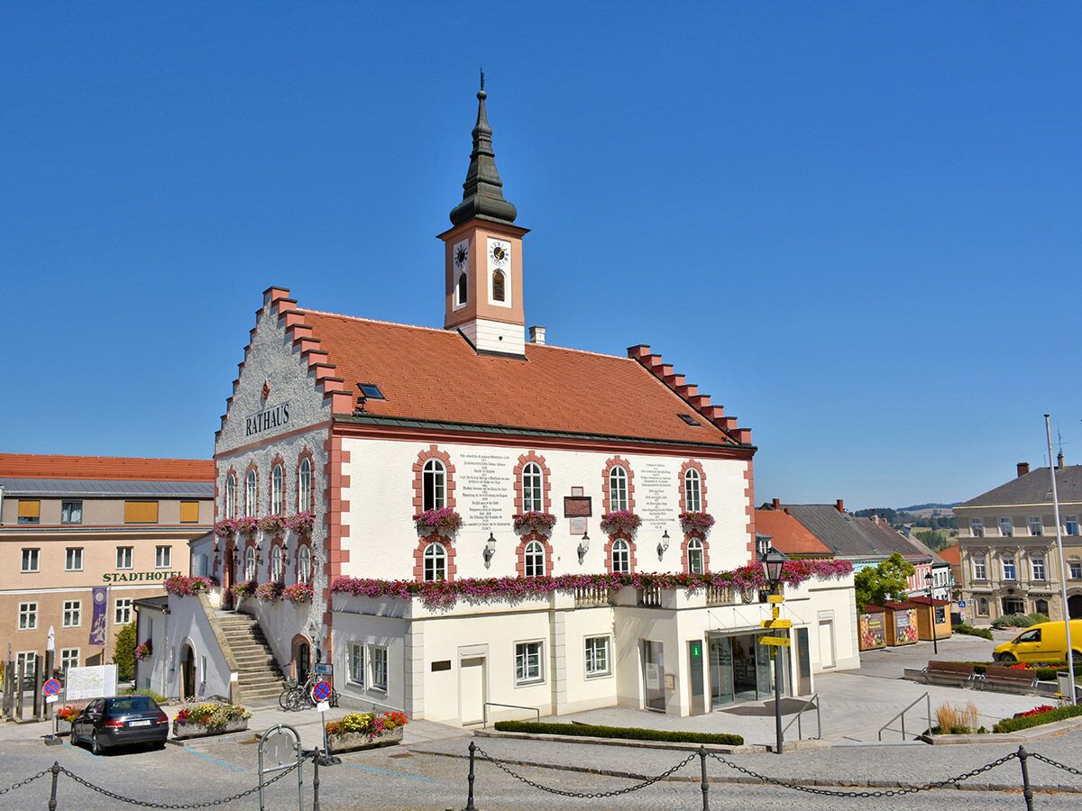 Historisches Rathaus mit Uhrturm, externer Steintreppe und üppigen bunten Blumenkästen unter blauem Himmel.