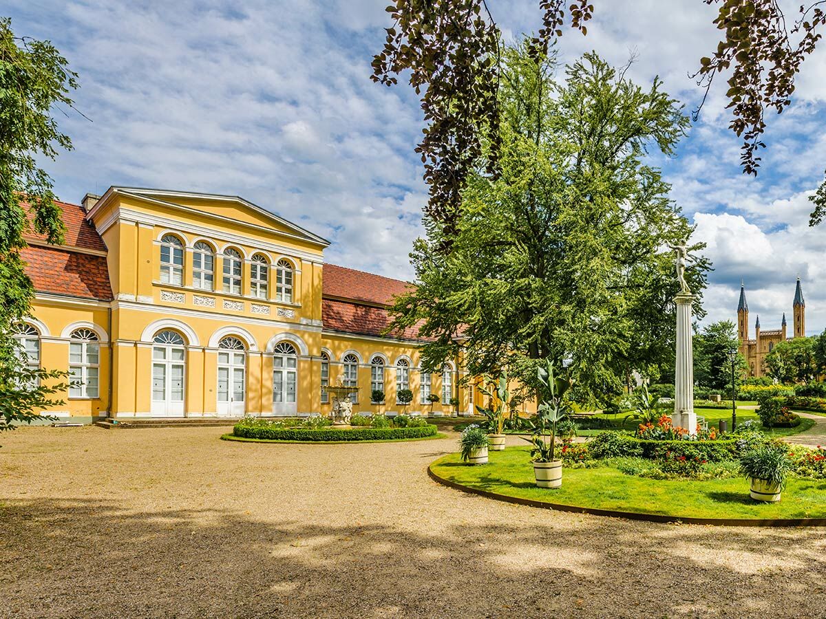 Gelbes historisches Schloss mit rotem Dach und Rundbogenfenstern, umgeben von einem grünen Garten, Statue, Bäumen und Türmen.