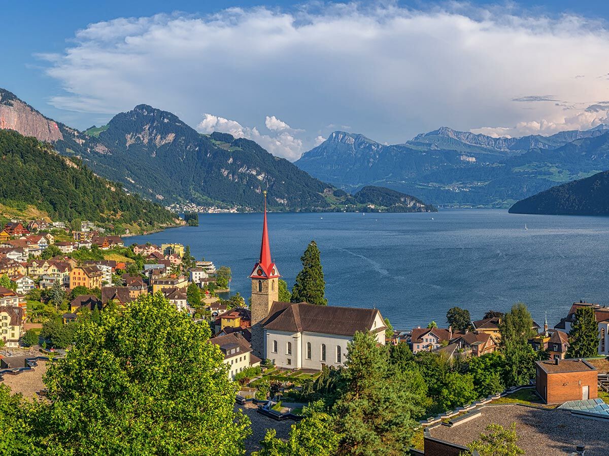 Blick auf malerisches Dorf mit roter Kirche am See, umgeben von grünen Bergen unter blauem Himmel in den Alpen.