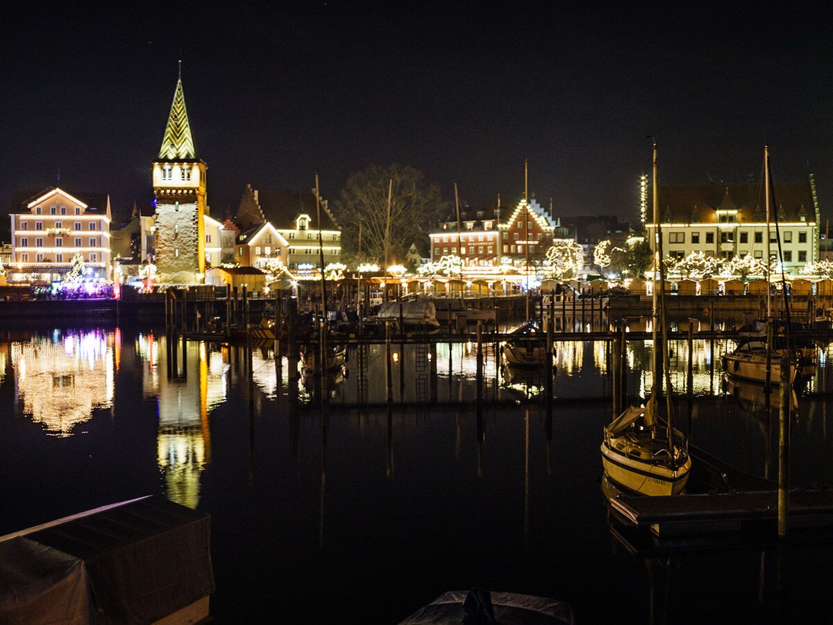 Festlicher Weihnachtsmarkt am Hafen von Lindau am Bodensee bei Nacht, mit beleuchtetem Mangturm, Gebäuden und Booten im Wasser.