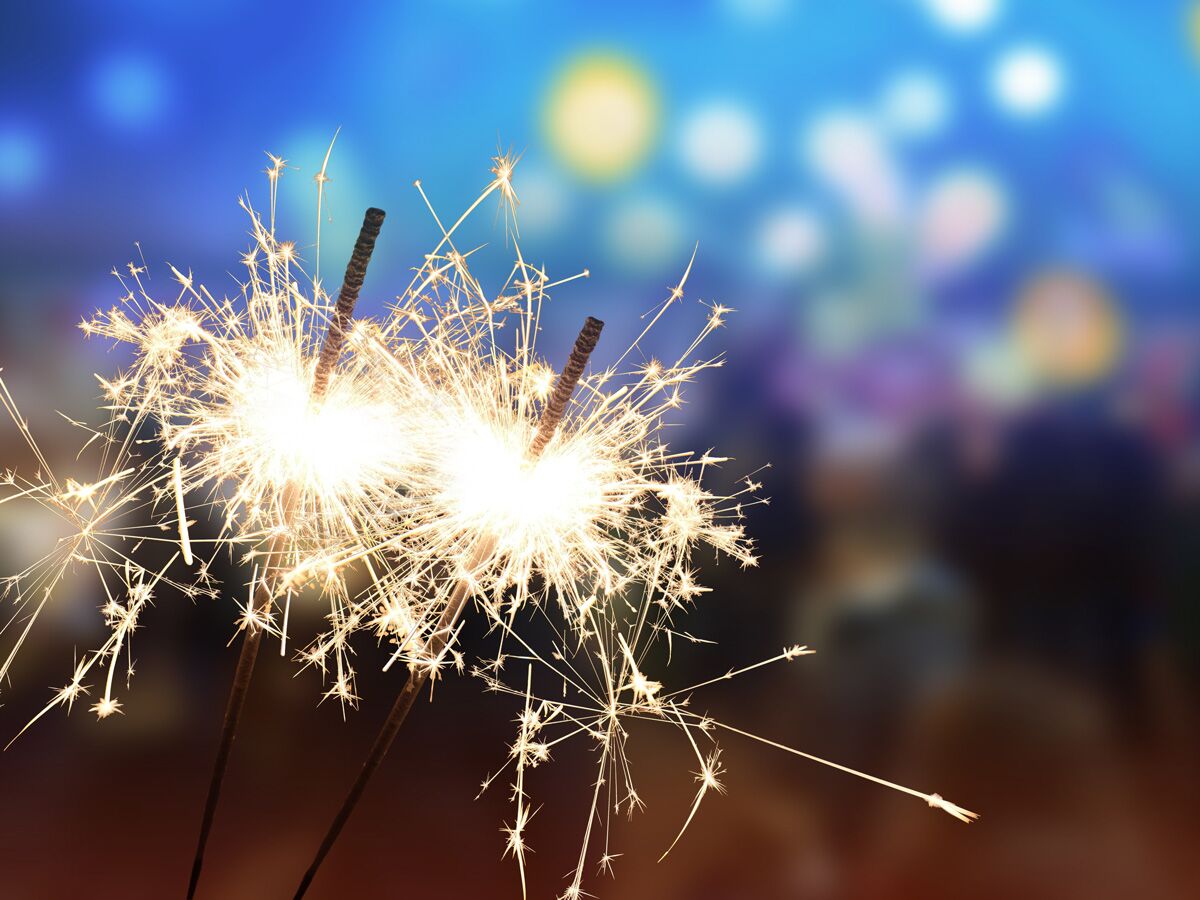 Nahaufnahme von brennenden Wunderkerzen, die helle Funken sprühen, vor einem verschwommenen Festlichter-Hintergrund.