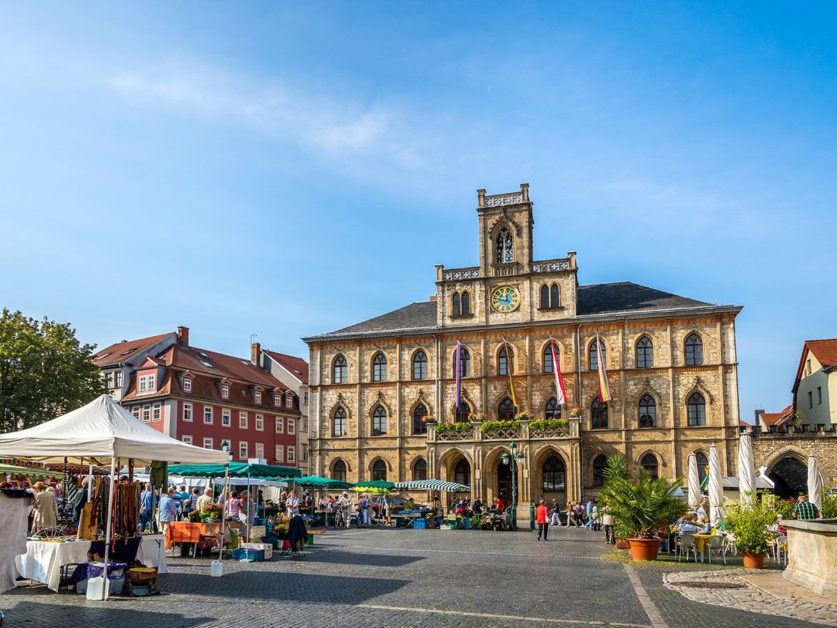 Belebter Marktplatz vor einem historischen Rathaus mit Turm und Uhr, umgeben von Marktständen und Cafés unter blauem Himmel.