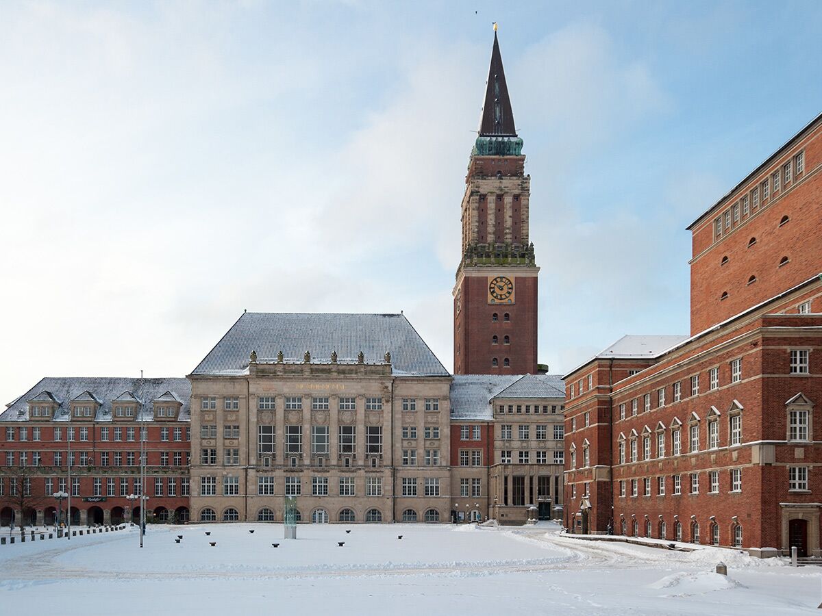 Historisches Kieler Rathaus mit Uhr-Turm und verschneitem Rathausplatz an einem Wintertag. Backstein- und Sandsteinfassade unter blauem Himmel.