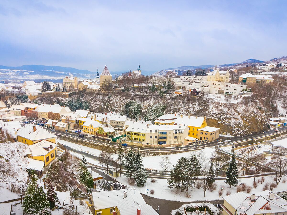 Panoramablick auf eine verschneite historische Stadt im Tal mit bunten Häusern, Kirche und Turm unter winterlichem Himmel.