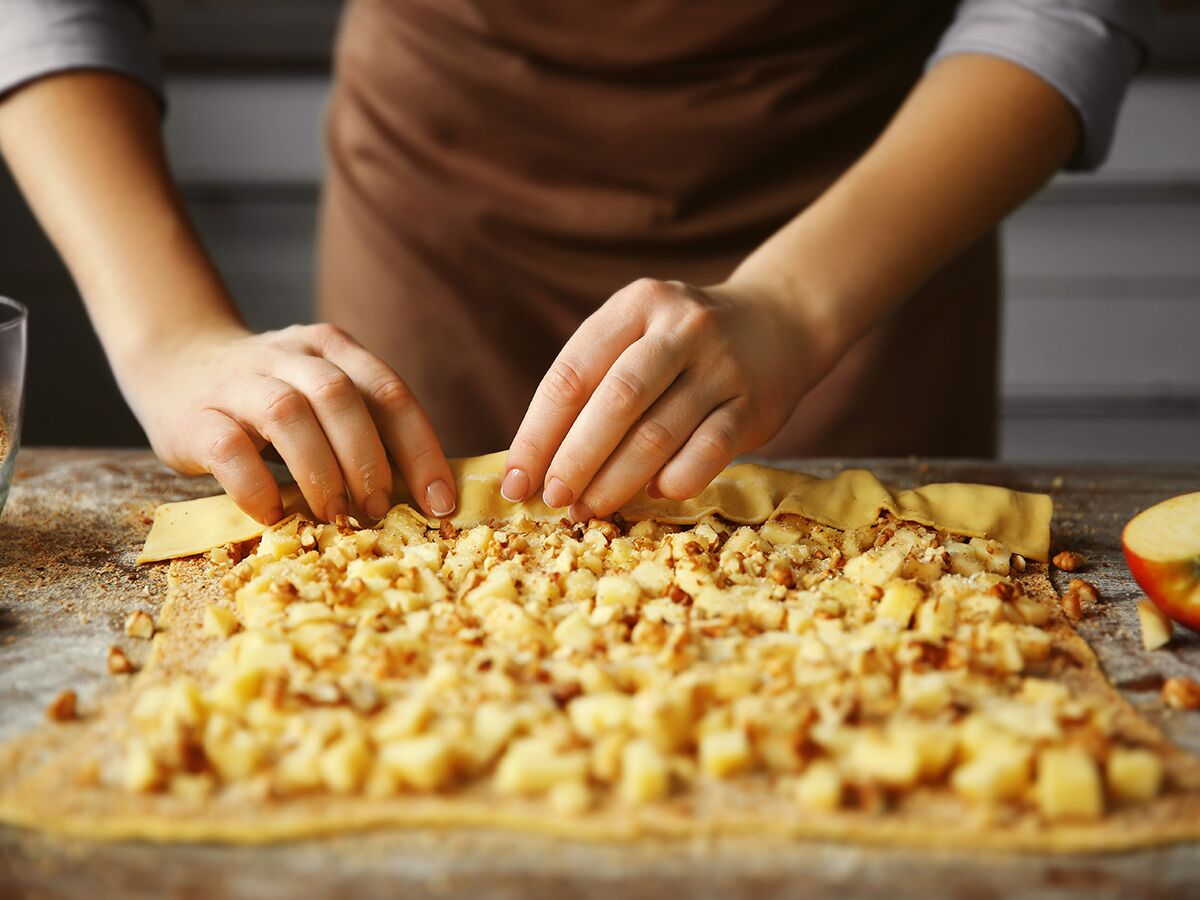 Nahaufnahme von Händen, die Apfelstrudel-Teig mit Apfel-Walnuss-Füllung auf einer Holzoberfläche einrollen.