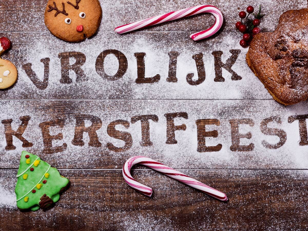 Weihnachtliche Plätzchen, Zuckerstangen und Beeren auf Holzhintergrund. „Vrolijk Kerstfeest“ in Puderzucker geschrieben.