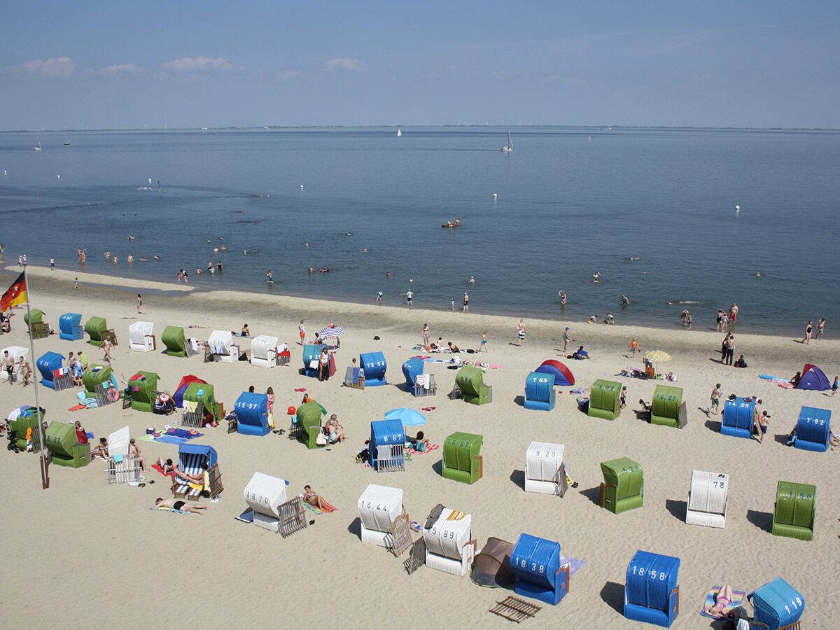 Luftaufnahme eines belebten deutschen Sandstrandes mit vielen bunten Strandkörben, badenden Menschen im Meer und Booten am Horizont unter blauem Himmel.