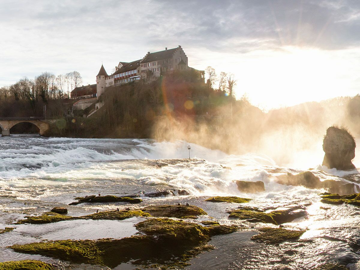 Majestätischer Rheinfall mit Schloss Laufen, leuchtendem Sonnenlicht und Wasserdunst, Felsen im Vordergrund.