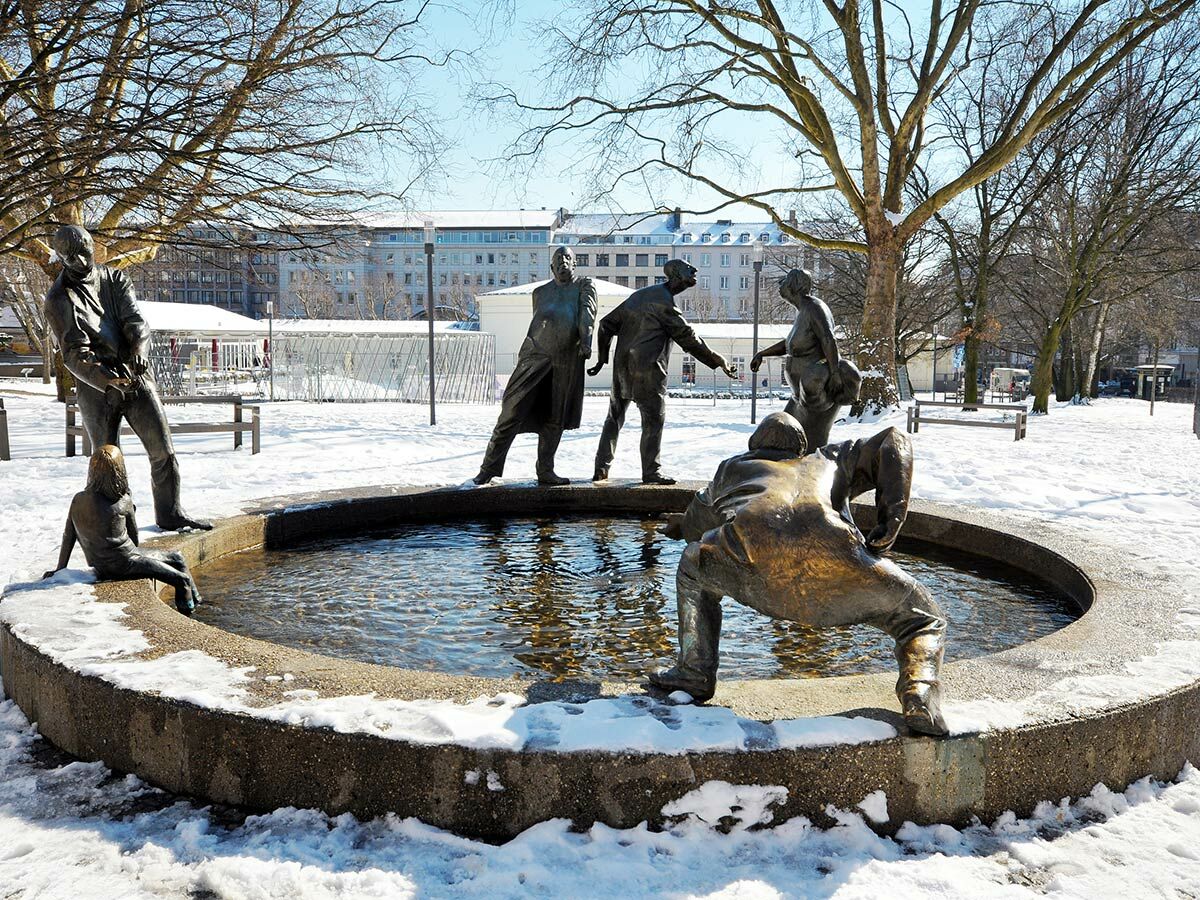 Bronzeskulptur-Brunnen mit mehreren lebensgroßen Figuren, die im Schnee in einem Berliner Park mit Wasser interagieren.