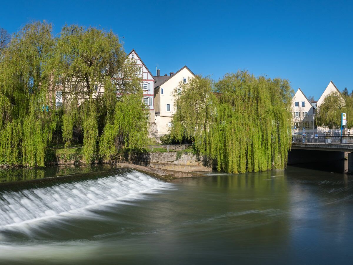 Langzeitbelichtung eines Flusswehrs mit grünen Weidenbäumen, traditionellen Häusern und blauem Himmel an einem sonnigen Tag.