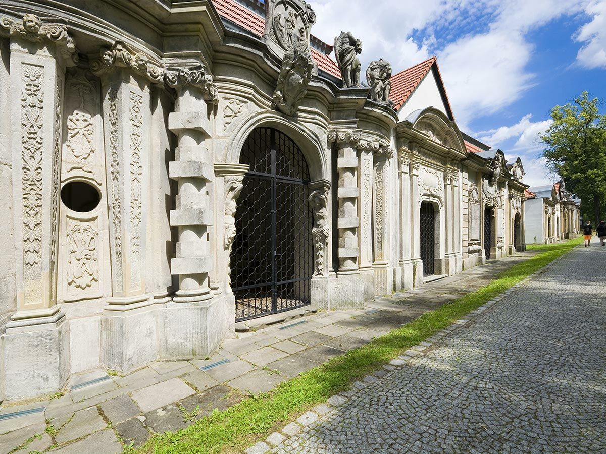 Langer, kunstvoll verzierter Barockbau aus hellem Stein mit gewölbten Eingängen und Skulpturen unter blauem Himmel.