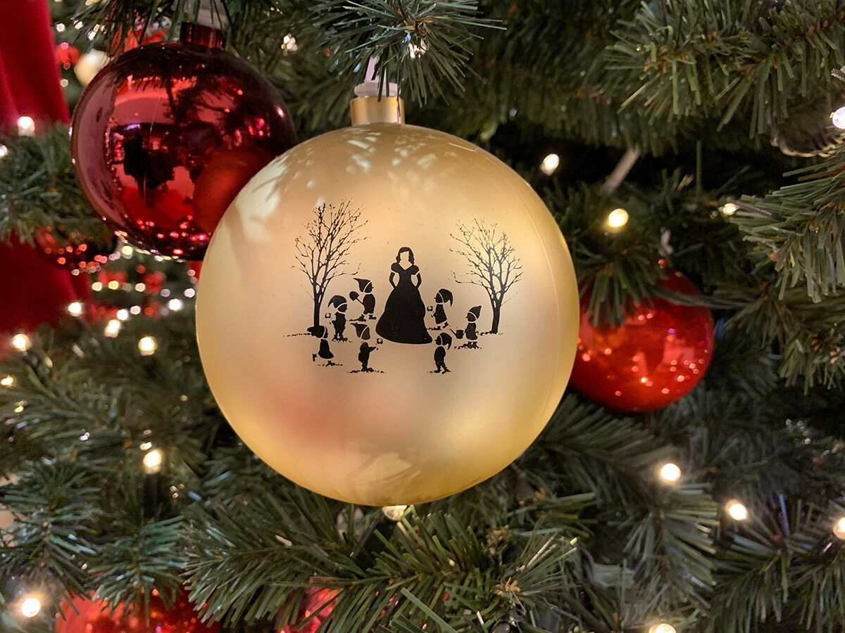 Goldene Christbaumkugel am Weihnachtsbaum mit Schneewittchen- und Zwerge-Silhouette, umgeben von roten Kugeln und Lichtern.