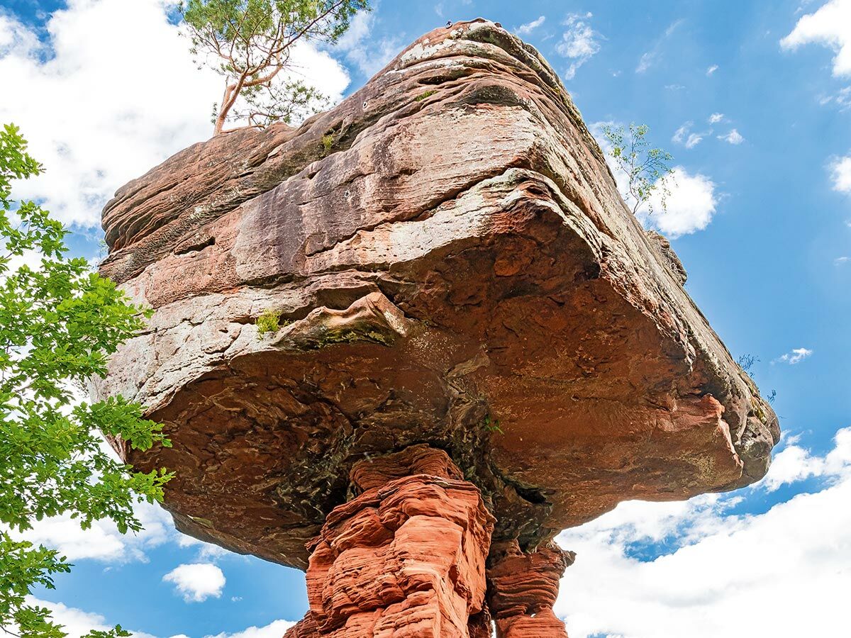 Einzigartige Balancing Rock Felsformation aus rotem Sandstein unter blauem Himmel mit weißen Wolken und grünen Bäumen.