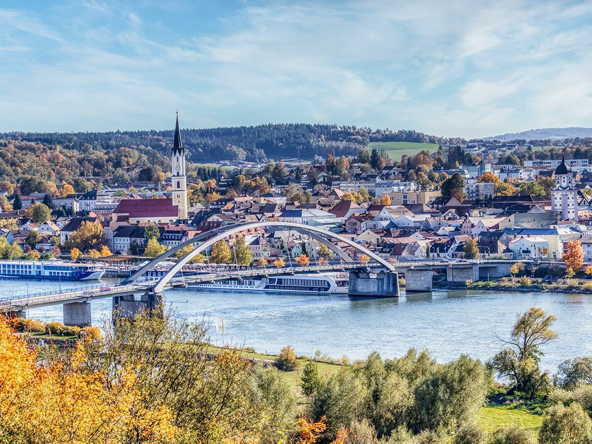Panorama von Vilshofen an der Donau im Herbst mit Fluss, Brücke, Kirche und bunten Bäumen unter blauem Himmel.