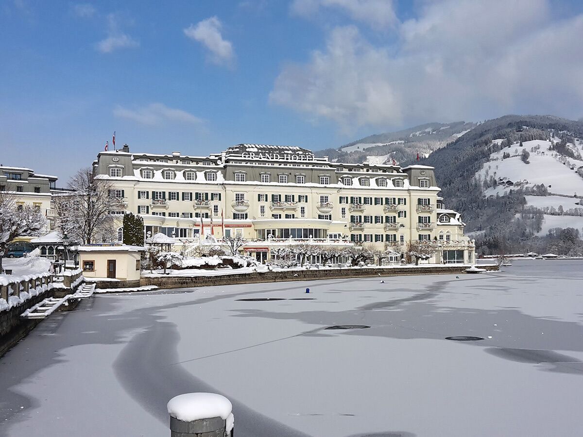 Luxuriöses Grand Hotel Zell am See am zugefrorenen Seeufer, umgeben von schneebedeckten Bergen unter blauem Himmel.