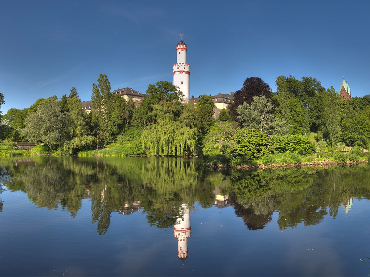 Majestätischer Wasserturm Bad Homburg mit rotem Dach, umgeben von grünen Bäumen, spiegelt sich im blauen Wasser des Schlossparks.