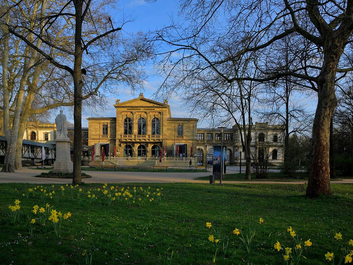 Kurhaus Baden-Baden mit blühenden Narzissen im Park, umgeben von Bäumen und einer Statue, am sonnigen Frühlingstag.