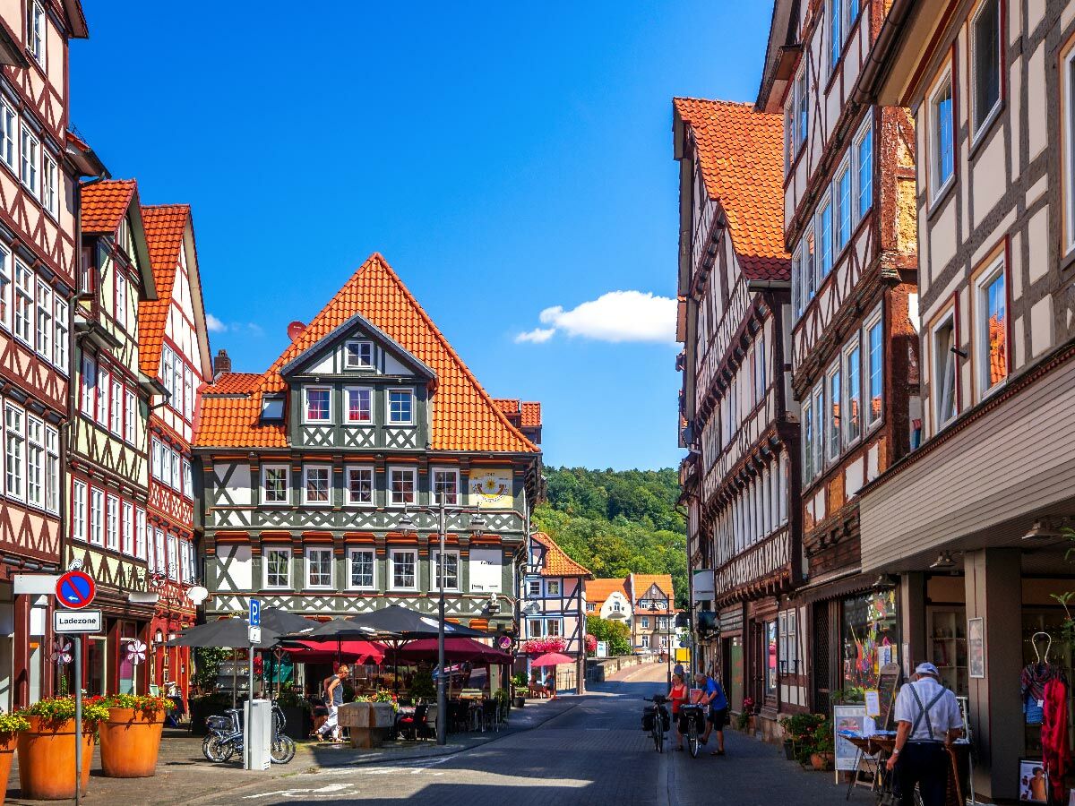 Belebte Gasse mit bunten Fachwerkhäusern und orangen Dächern unter blauem Himmel in einer deutschen Altstadt mit Cafés und Passanten.