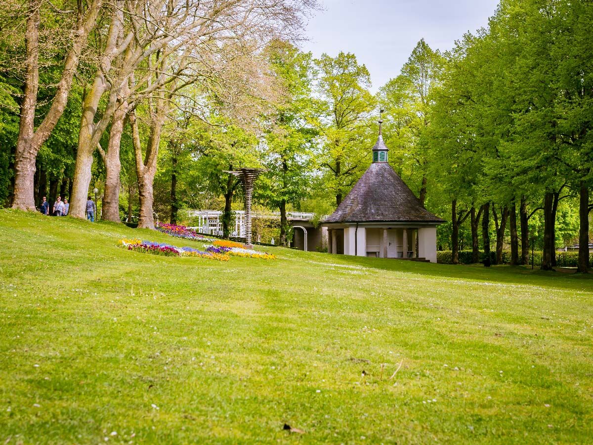 Grüner Park mit historischem Pavillon, bunten Blumenbeeten und Spaziergängern unter Bäumen an einem sonnigen Tag.