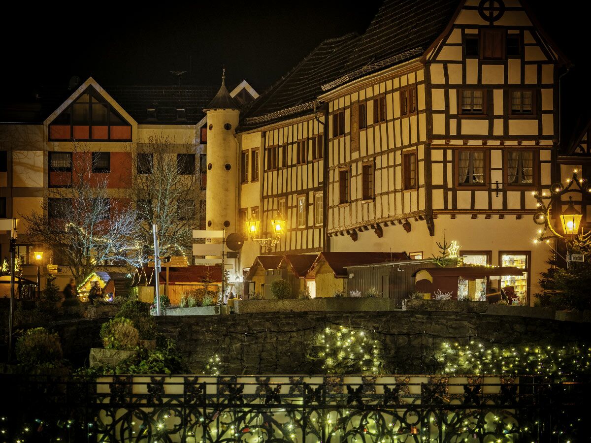 Festlicher Weihnachtsmarkt in einer europäischen Stadt bei Nacht mit beleuchteten Fachwerkhäusern, Bäumen und einer Brücke mit Lichterketten.