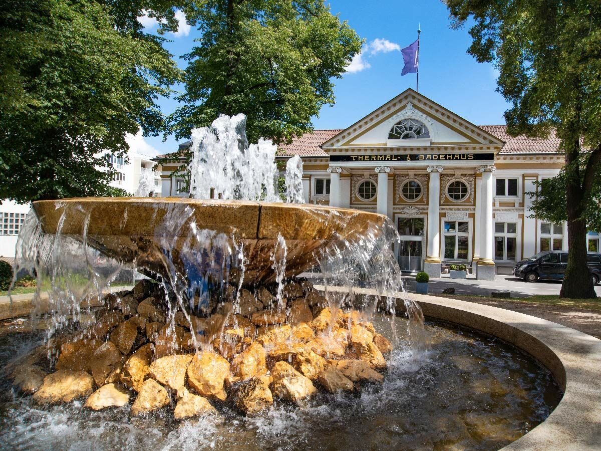 Großer Steinbrunnen mit Wasser vor dem Thermal- &amp; Badehaus. Elegantes Gebäude mit Säulen, goldener Verzierung, Bäume, blauer Himmel.