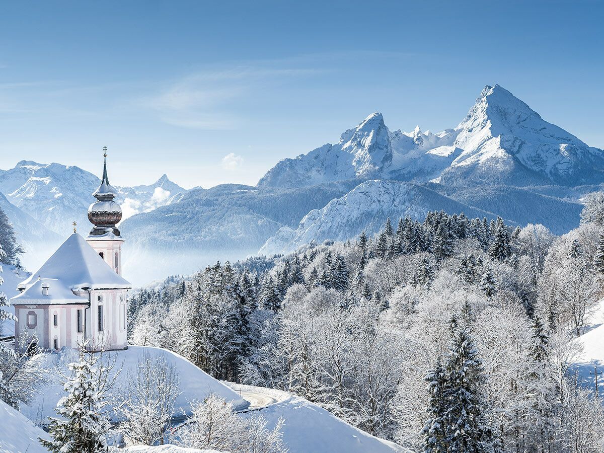 St. Sebastian Kirche in Ramsau, verschneite Berge und Wälder in winterlicher Alpenlandschaft bei blauem Himmel.