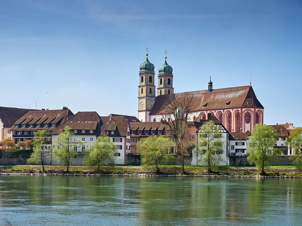 Panoramablick auf bayerische Altstadt mit markanter Kirche und bunten Häusern entlang eines Flusses unter blauem Himmel im Frühling.