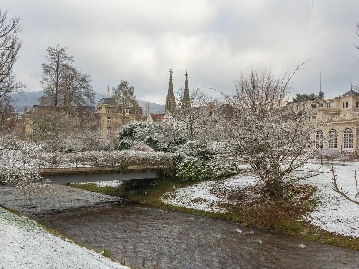 Schneebedeckte Winterlandschaft mit Fluss Oos, Brücke, kahlen Bäumen und historischen Gebäuden in Baden-Baden, Deutschland.