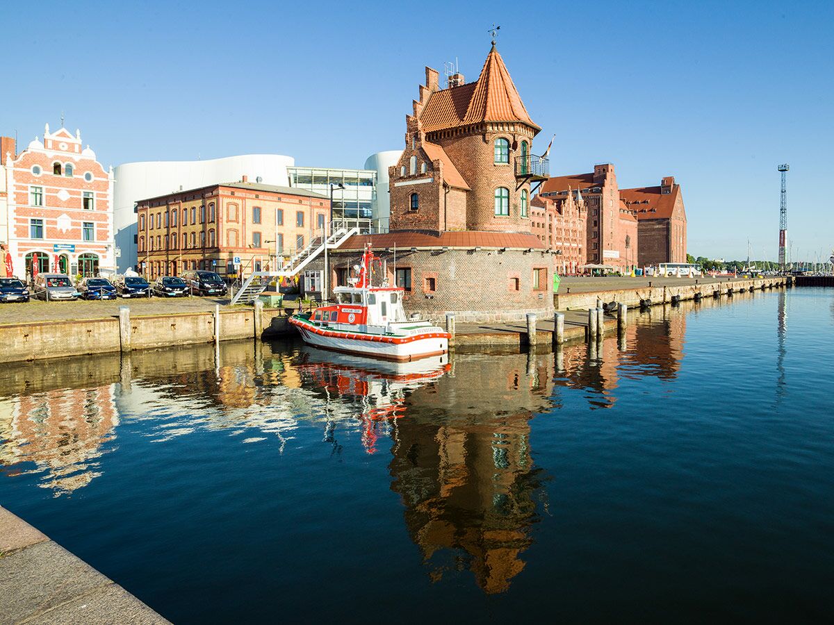 Blick auf den historischen Hafen von Stralsund mit Backsteingebäuden, modernem Museum und einem roten Lotsenboot bei Sonnenschein.