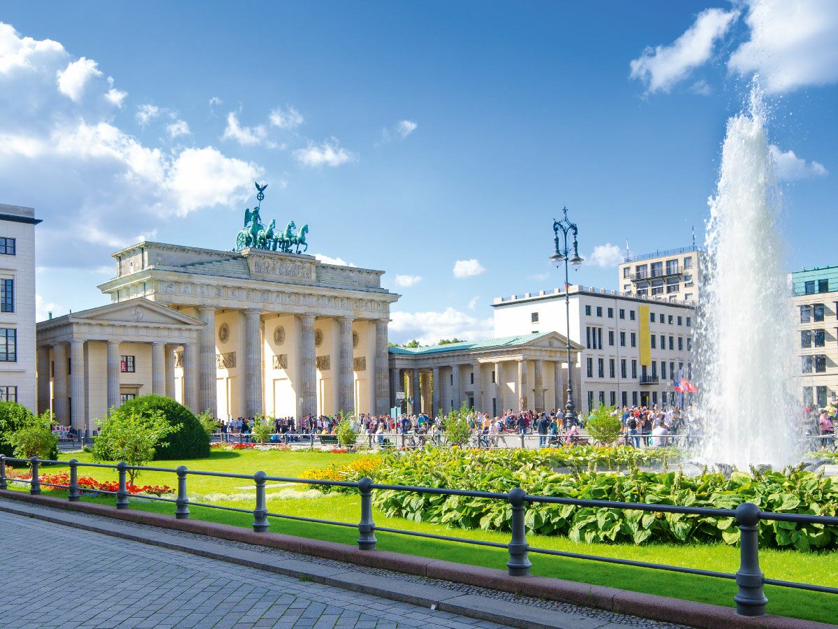 Das Brandenburger Tor in Berlin bei sonnigem Wetter mit der Quadriga, einem Springbrunnen und vielen Menschen.