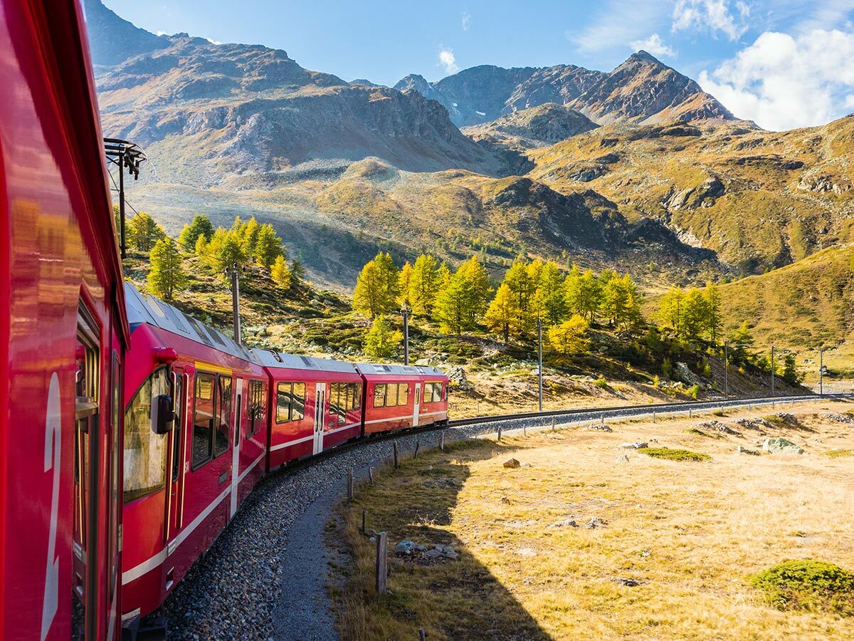 Ein roter Zug fährt auf einer kurvigen Bahnstrecke durch eine herbstliche Alpenlandschaft mit gelben Bäumen und Bergen unter blauem Himmel.