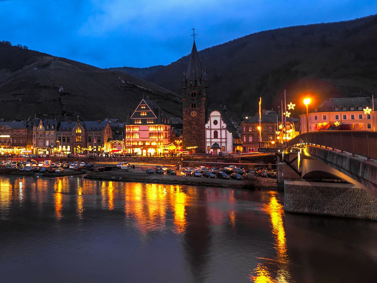 Cochem an der Mosel bei Nacht mit beleuchteten Gebäuden, Fluss und Brücke. Malerische Stadtansicht mit Weinbergen.