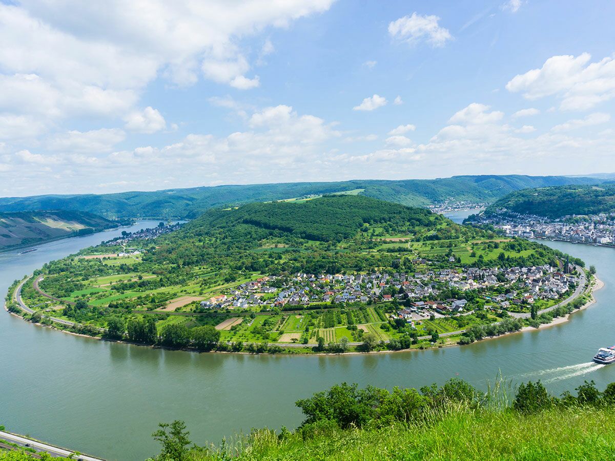 Luftaufnahme der Bopparder Rheinschleife mit malerischer Stadt, Fluss, grünen Hügeln, Wäldern, Straße und Schiff unter blauem Himmel.
