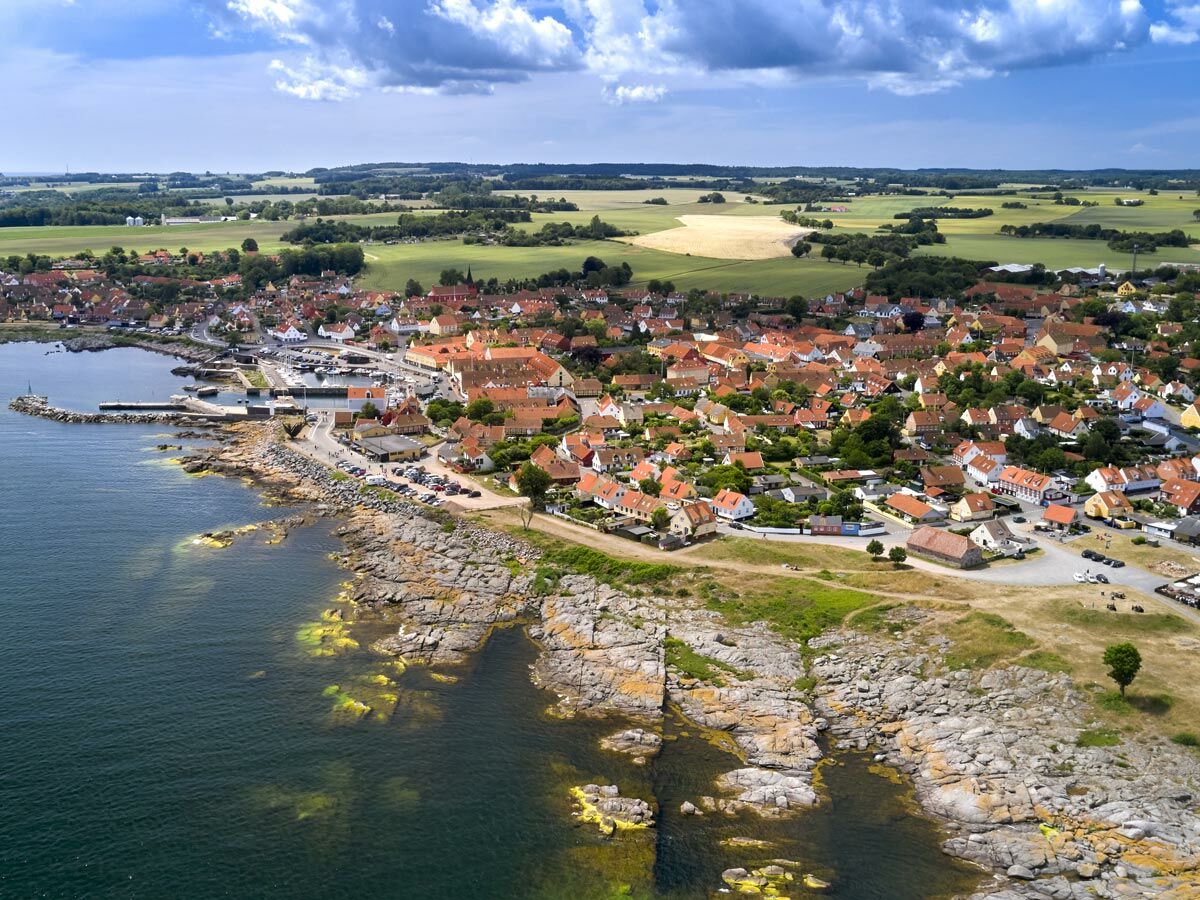 Luftaufnahme einer malerischen Küstenstadt mit roten Dächern, einem geschäftigen Hafen, felsiger Küste und weiten Feldern im Hintergrund.