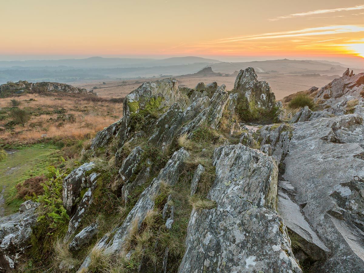 Weitblick über eine felsige Moorlandschaft bei Sonnenuntergang mit orangerotem Himmel und entfernten Hügeln.