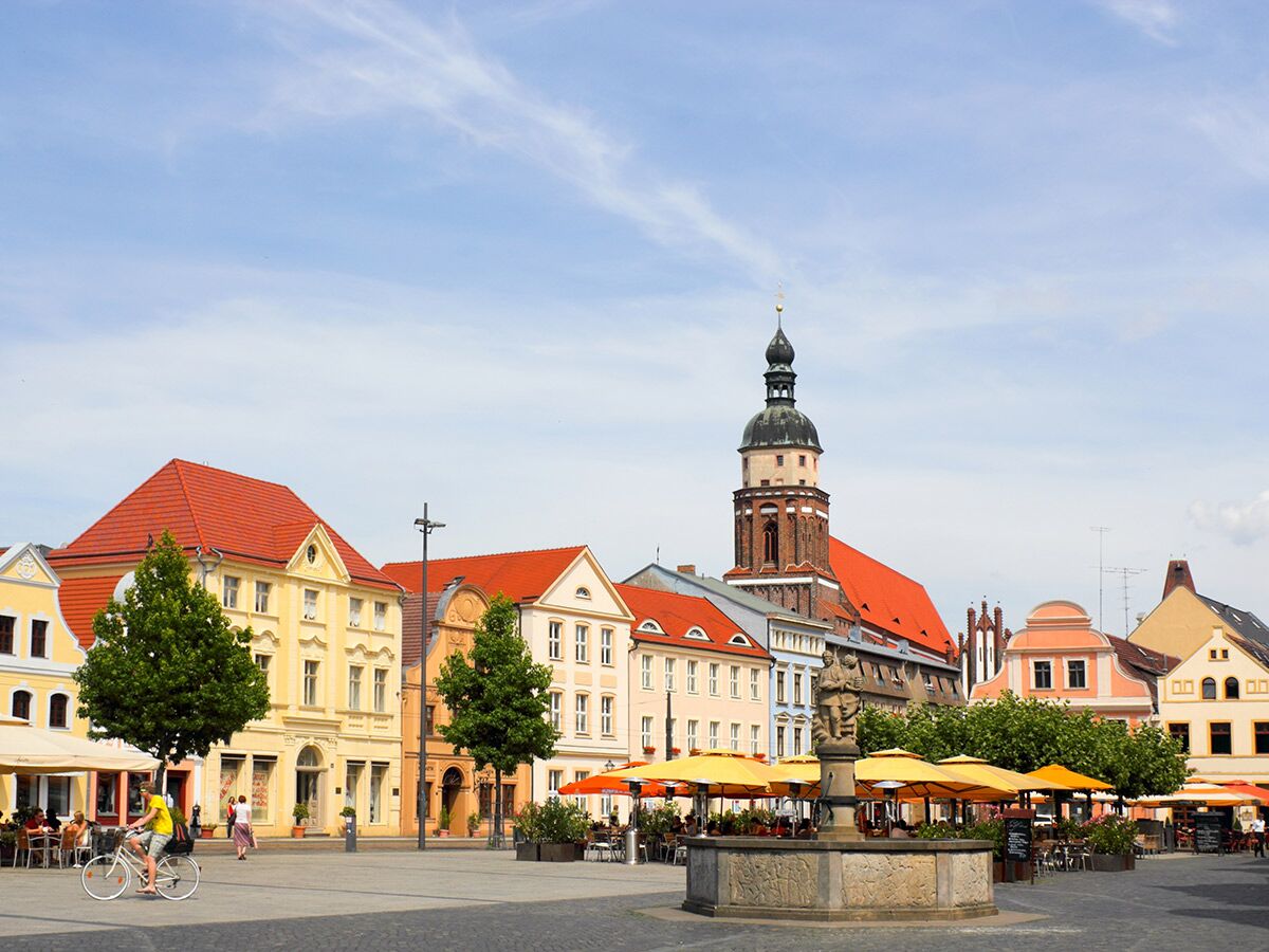 Sonniger historischer Marktplatz in Görlitz mit farbigen Gebäuden, Kirchenturm, Brunnen, belebten Außencafés und Passanten.