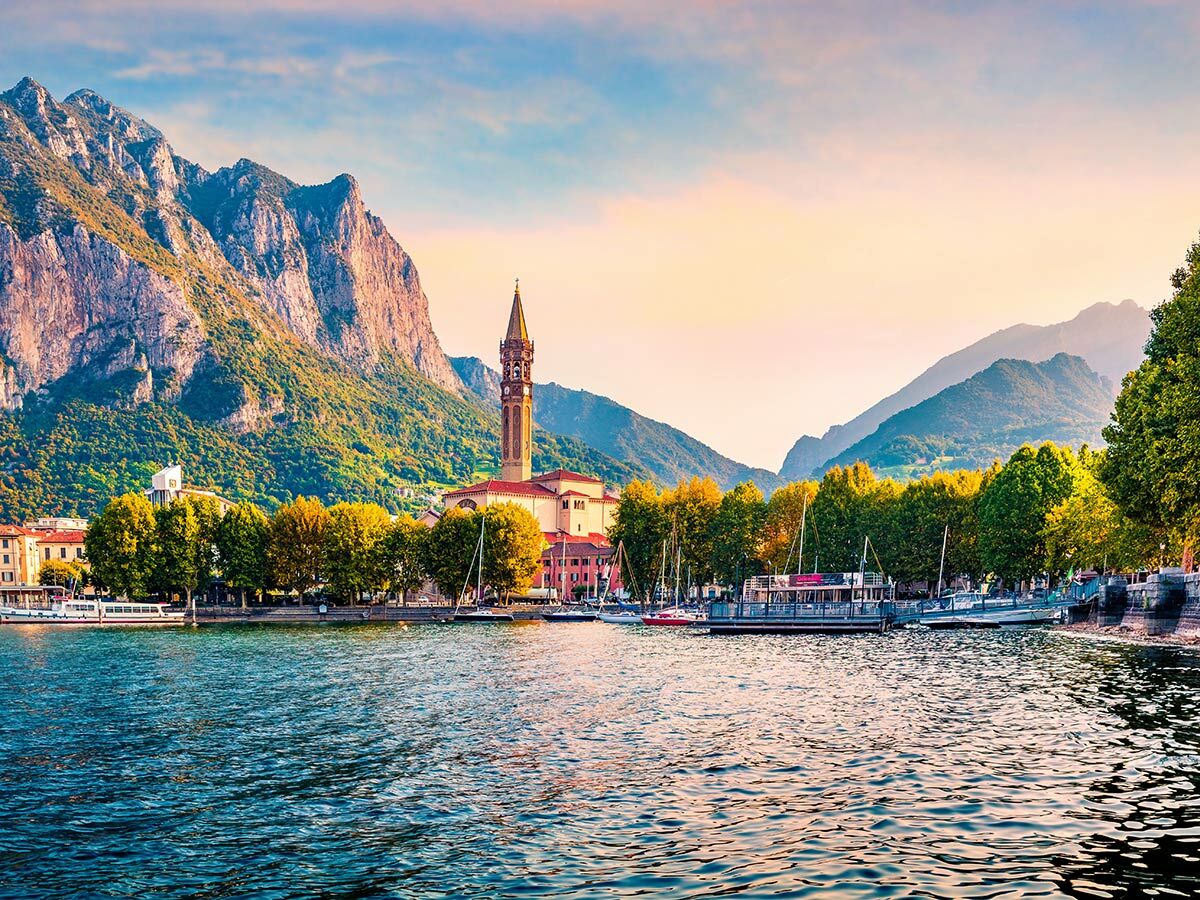 Malerischer Blick auf Lecco am Comer See mit markantem Glockenturm, Booten, Uferpromenade und hohen Bergen bei Sonnenuntergang.