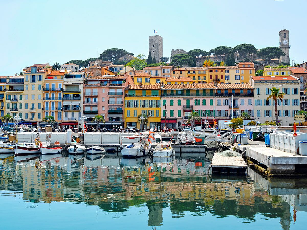 Panoramablick auf den Hafen von Cannes mit bunten Häusern, Booten, Altstadt und einem Glockenturm unter blauem Himmel.