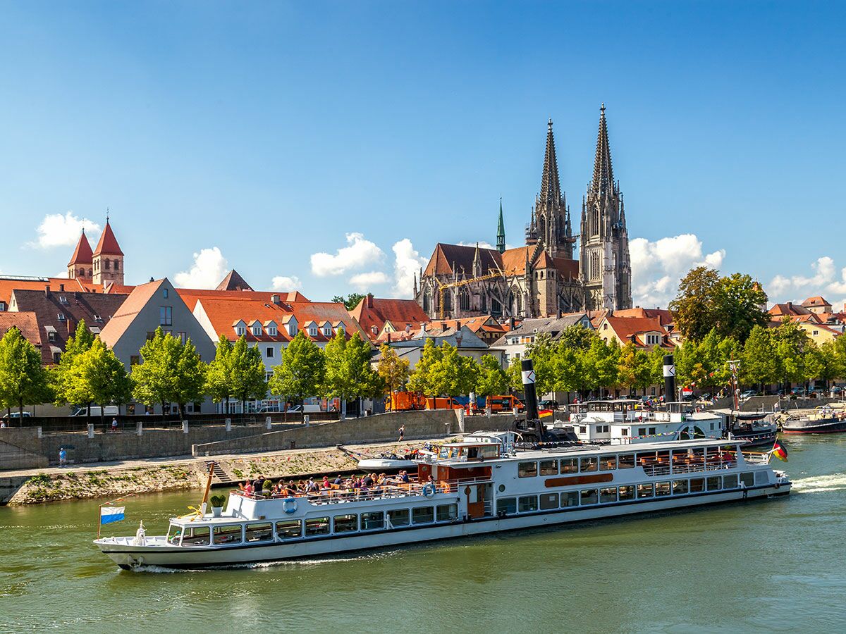 Panoramablick auf Regensburg mit Donau, fahrendem Ausflugsschiff voller Touristen und Dom St. Peter unter blauem Himmel.