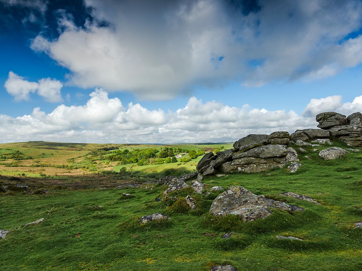 Weite, grüne Moorlandschaft mit großen Felsformationen im Vordergrund unter einem blauen Himmel mit weißen Wolken.